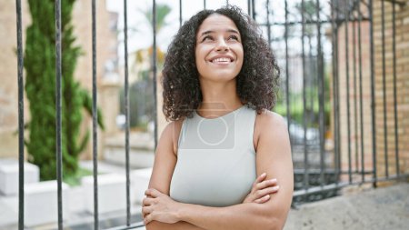 Fröhliche junge hispanische Frau mit ihren schönen lockigen Haaren und ihrem ansteckenden Lächeln, die draußen im Herzen der Stadt steht und Positivität ausstrahlt, die Arme in einer selbstbewussten Haltung verschränkt