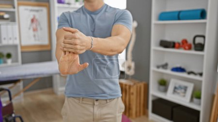 Photo for A young hispanic man stretching his wrist in a well-equipped rehabilitation clinic. - Royalty Free Image