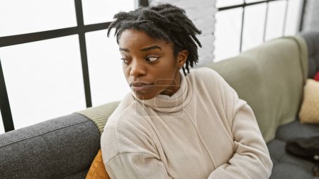 Photo for African american woman with dreadlocks sitting thoughtfully on a sofa in a modern living room. - Royalty Free Image