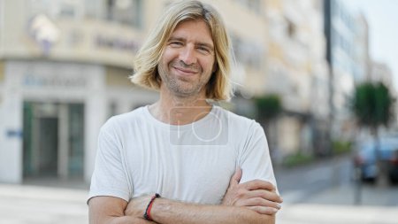 Schöner lächelnder Mann mit langen blonden Haaren steht selbstbewusst auf einer sonnigen urbanen Straße.