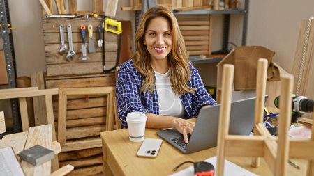A smiling woman engages in business on a laptop at a carpentry workshop surrounded by tools and woodwork.