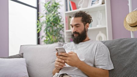 Téléchargez les photos : Bel homme hispanique avec barbe en utilisant smartphone sur canapé confortable dans un salon intérieur moderne - en image libre de droit
