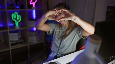 Blonde woman making heart shape with hands in a neon-lit gaming room at night
