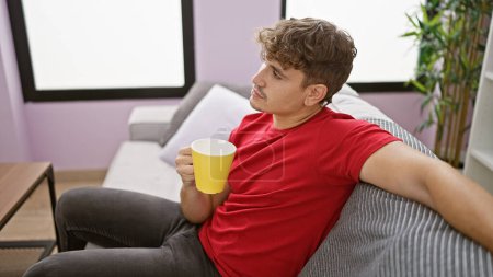 Entspannter Morgen mit einer Tasse Kaffee, Porträt eines jungen hispanischen Mannes, der es sich zu Hause auf seinem Wohnzimmersofa bequem macht