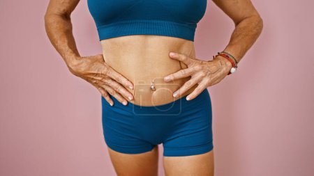 Photo for A fit middle-aged woman touches her toned abdomen against a pink background, implying health and wellness. - Royalty Free Image