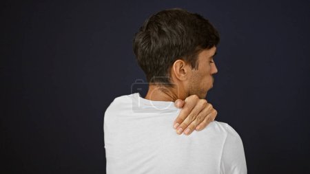 Photo for Distressed young hispanic man suffering intense back pain, isolated on a black background, touching an injury while looking away with a concentrated and serious expression - Royalty Free Image