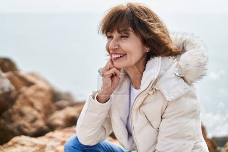 Téléchargez les photos : Femme d'âge moyen souriant confiant assis sur le rocher au bord de la mer - en image libre de droit