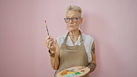 Passionate senior grey-haired woman artist in apron and glasses, wielding paintbrush over canvas and palette, speaks art lesson isolated on pink background