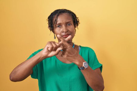 Foto de Mujer africana con rastas de pie sobre fondo amarillo expresión de rechazo cruzando dedos haciendo signo negativo - Imagen libre de derechos