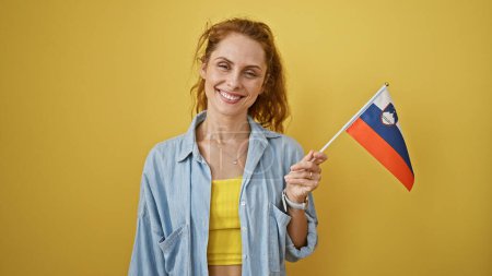 Photo for Smiling young caucasian woman holding a slovenian flag against a solid yellow background, exuding charm and positivity. - Royalty Free Image