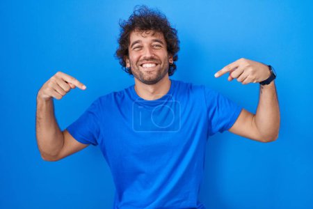 Photo for Hispanic young man standing over blue background looking confident with smile on face, pointing oneself with fingers proud and happy. - Royalty Free Image