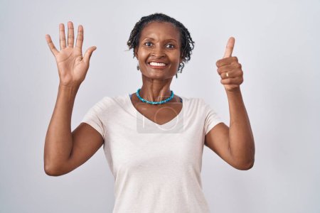 Foto de Mujer africana con rastas de pie sobre fondo blanco mostrando y apuntando hacia arriba con los dedos número seis mientras sonríe confiado y feliz. - Imagen libre de derechos