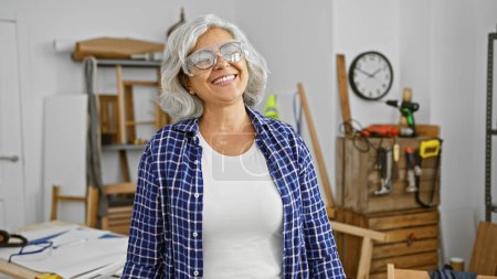 Téléchargez les photos : Une femme joyeuse d'âge moyen aux cheveux gris portant des lunettes de sécurité dans un atelier de menuiserie. - en image libre de droit