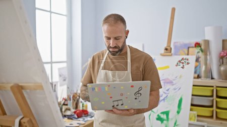 Photo for Handsome young man artist, relaxed yet concentrated, expertly handling his laptop amidst paintbrushes and canvases in bustling art studio - Royalty Free Image