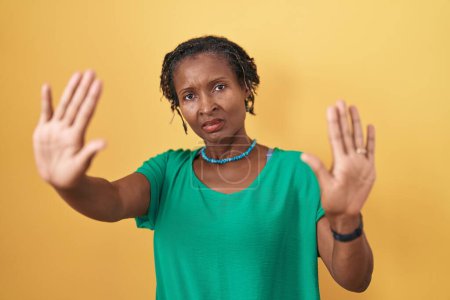 Foto de Mujer africana con rastas de pie sobre fondo amarillo haciendo marco usando las manos palmas y los dedos, perspectiva de la cámara - Imagen libre de derechos