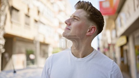 Schöner junger kaukasischer Mann, der auf einer belebten Straße der Stadt steht, aufblickt, selbstbewusst lächelt, Freude in den Himmel ausstrahlt und in seinem lässigen Look einen coolen, positiven Lebensstil widerspiegelt.