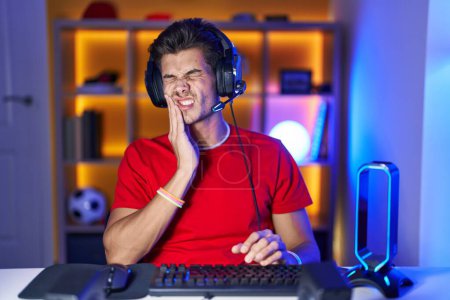 Téléchargez les photos : Jeune homme hispanique jouant à des jeux vidéo touchant la bouche avec la main avec une expression douloureuse à cause de maux de dents ou de maladies dentaires sur les dents. dentiste - en image libre de droit