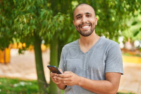 Foto de Joven latino sonriendo confiado usando smartphone en el parque - Imagen libre de derechos