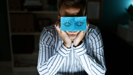 A humorous portrait of a young man in an office, holding a sticky note with drawn eyes over his face.
