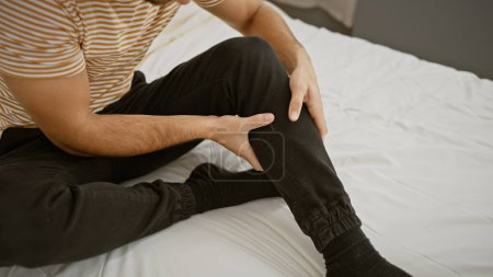 Photo for Handsome young hispanic man in casual attire touching his leg in pain, sitting on a white bed indoors. - Royalty Free Image