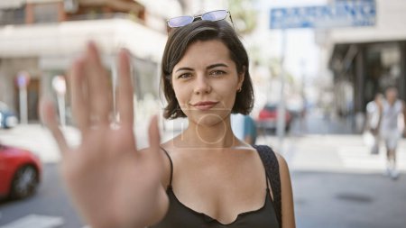 Mujer hispana joven enojada, con una expresión hermosa pero seria, ordena enérgicamente '¡para!' con su mano al sol en la calle de la ciudad.