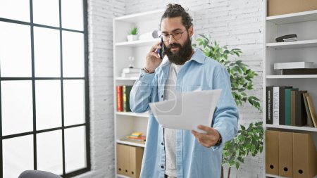 Photo for Handsome hispanic man with beard talking on phone and reading document in modern office setting. - Royalty Free Image