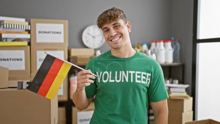Foto de Retrato conmovedor, joven hispano sonriente, un voluntario dedicado en un centro de caridad, orgullosamente sosteniendo la bandera alemana - Imagen libre de derechos