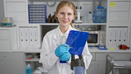 Téléchargez les photos : Une scientifique blonde tenant un presse-papiers dans un laboratoire moderne, transmettant expertise et professionnalisme. - en image libre de droit