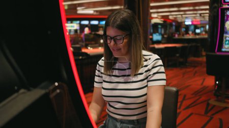 Foto de Mujer hispana adulta sonriendo mientras juega a la máquina tragaperras en un vibrante entorno de casino, que representa el ocio y el juego. - Imagen libre de derechos