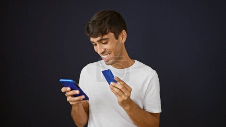 Photo for Confident young hispanic man happily typing a message, enjoying online shopping with credit card and smartphone on black isolated background - Royalty Free Image