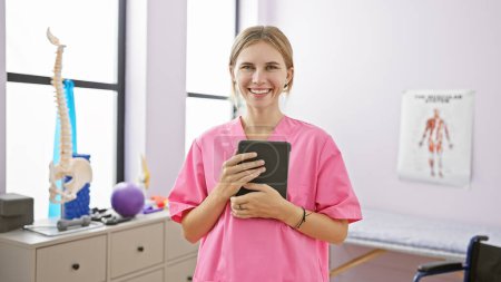 Foto de Mujer sonriente en matorrales rosados sosteniendo la tableta en una clínica de rehabilitación bien equipada con carteles anatómicos - Imagen libre de derechos