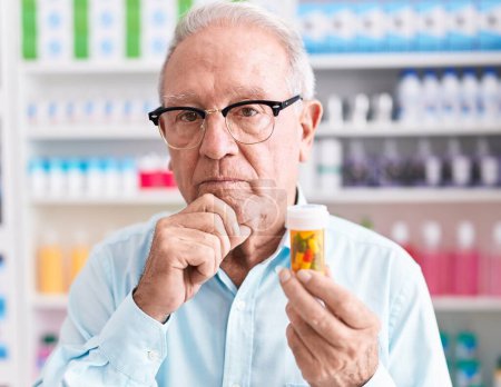 Téléchargez les photos : Homme âgé avec les cheveux gris tenant des pilules à la pharmacie visage sérieux penser à la question avec la main sur le menton, réfléchi à l'idée déroutante - en image libre de droit
