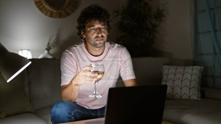 Photo for A thoughtful young man sits with a wine glass in a cozy living room at night, concentrating on a laptop screen. - Royalty Free Image