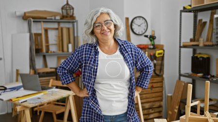 Confident grey-haired woman standing with arms crossed in a well-equipped carpentry workshop, projecting experience