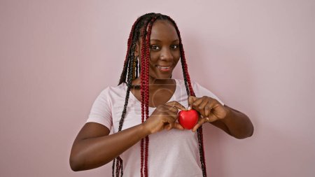 Téléchargez les photos : Belle femme afro-américaine exhibe son sourire confiant tout en tenant un cœur, suintant de joie sur le fond rose isolé - en image libre de droit