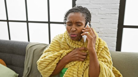 Photo for Mature african american woman speaking on phone, frowning, wrapped in yellow blanket, indoors - Royalty Free Image