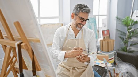 Photo for Confident, smiling young hispanic man with grey hair joyfully drawing his masterpiece in a cozy art studio - Royalty Free Image