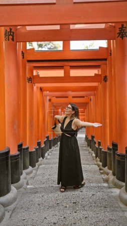 Téléchargez les photos : Belle femme hispanique, joyeuse à lunettes, embrasse la liberté, souriant autour des portes torii fushimi, les bras ouverts en altitude dans les beaux moments de la vie - en image libre de droit