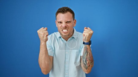 Téléchargez les photos : Jeune homme hispanique en colère avec les poings serrés de barbe contre le mur bleu. - en image libre de droit