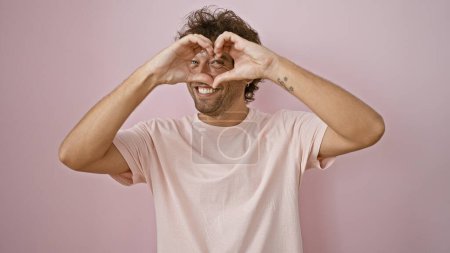 Photo for A cheerful young man making a heart symbol with his hands against a plain pink background. - Royalty Free Image