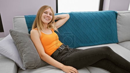 Téléchargez les photos : Femme blonde souriante détendue sur un canapé moderne dans un élégant salon intérieur. - en image libre de droit
