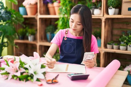 Photo for Young chinese woman florist counting dollars writing on notebook at flower shop - Royalty Free Image