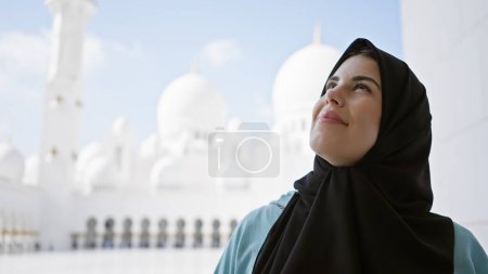 Photo for A young adult hispanic woman wearing a hijab looks contemplatively at the islamic architecture of a uae mosque against a bright sky. - Royalty Free Image