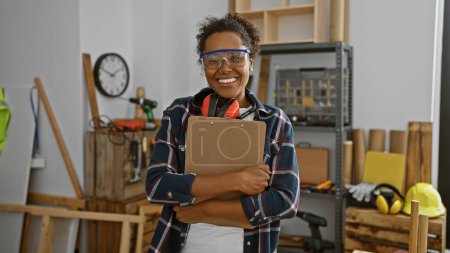 Foto de Mujer sonriente con gafas de seguridad y auriculares sosteniendo un portapapeles en un taller de carpintería - Imagen libre de derechos