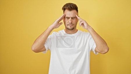 Ein junger hispanischer Mann mit Bart posiert vor gelbem Hintergrund und zeigt Stress oder Kopfschmerzen, während er ein weißes Hemd trägt.