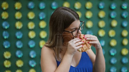 Beautiful hispanic woman in glasses delightfully eating traditional japanese melon pan snack on tokyo's vibrant street