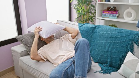 Téléchargez les photos : Jeune homme hispanique couché sur le canapé couvrant le visage avec un coussin à la maison - en image libre de droit