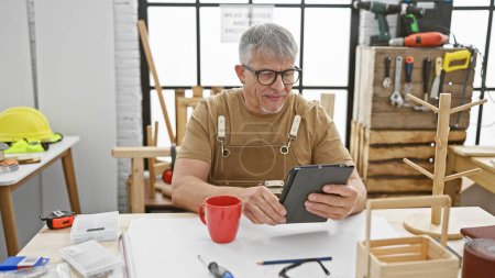 Foto de Un hombre de pelo gris sonriente usando una tableta en un taller de carpintería con herramientas y carpinterías visibles. - Imagen libre de derechos