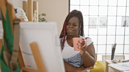 Thinking over a morning cup of coffee, african american artist woman draws intently at her art studio, looking at canvas with a mix of doubt and determination.
