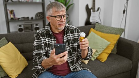 Foto de Hombre de mediana edad revisando la botella de medicamentos mientras usa el teléfono inteligente en la sala de estar. - Imagen libre de derechos